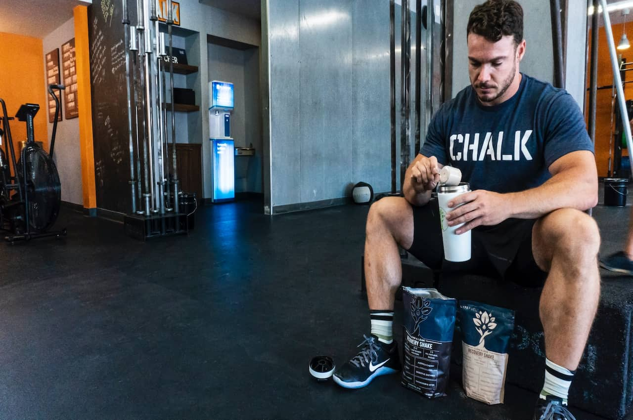 A man wearing a blue shirt and black shorts is using a brown scoop to put protein powder on a white tumbler