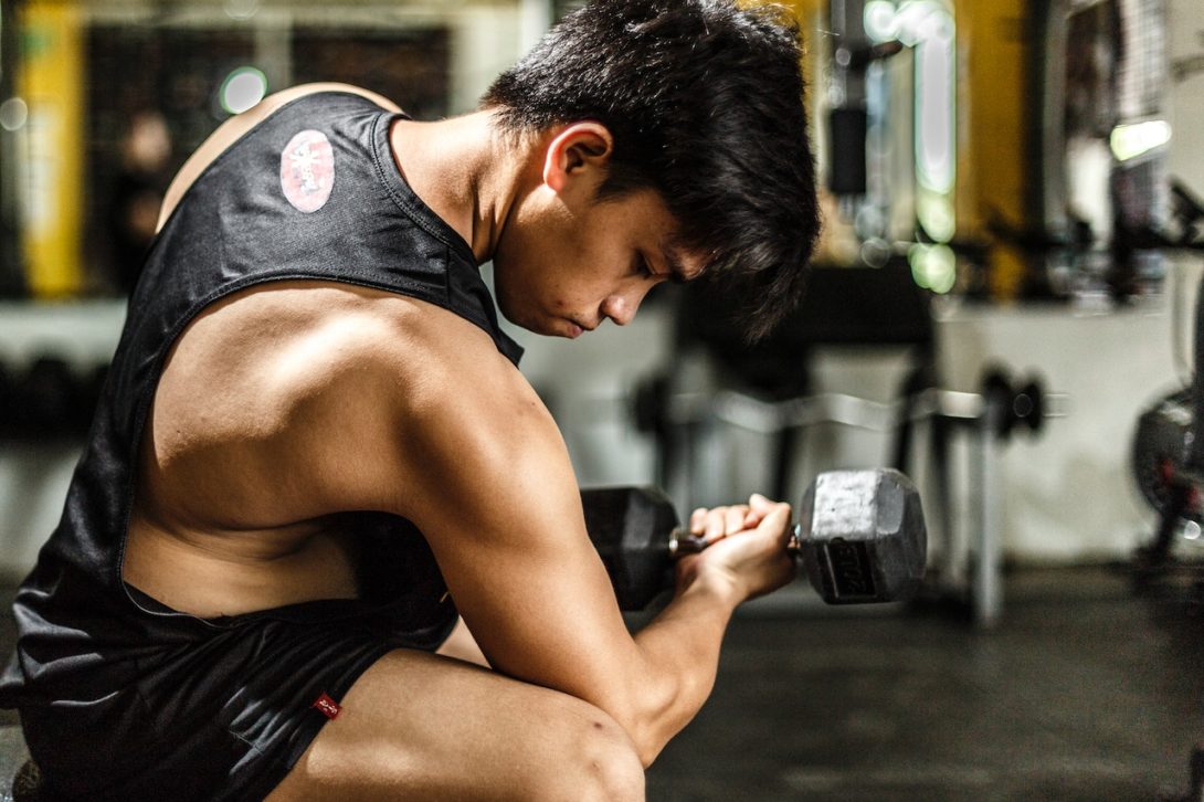 A man wearing a black tank top and black shorts is using a black dumbbell inside the gym