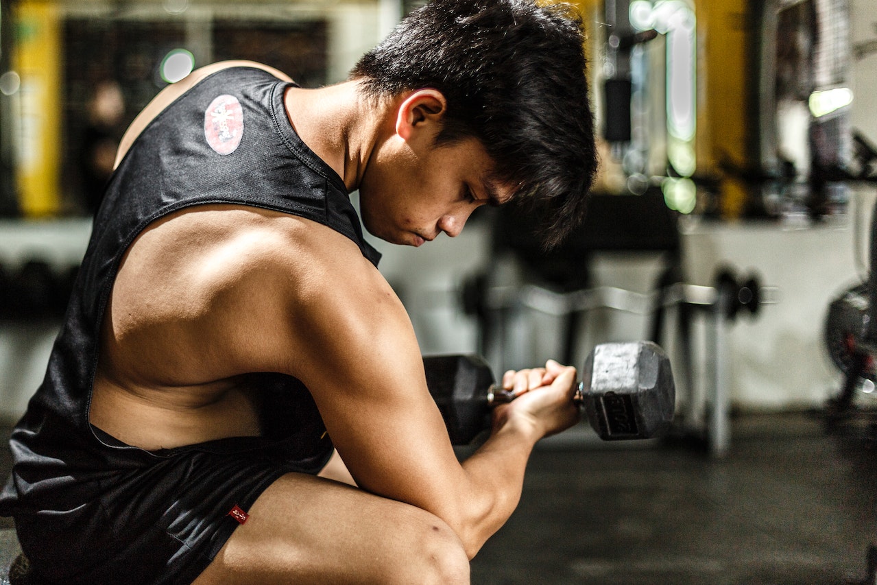 A man wearing a black tank top and black shorts is using a black dumbbell inside the gym