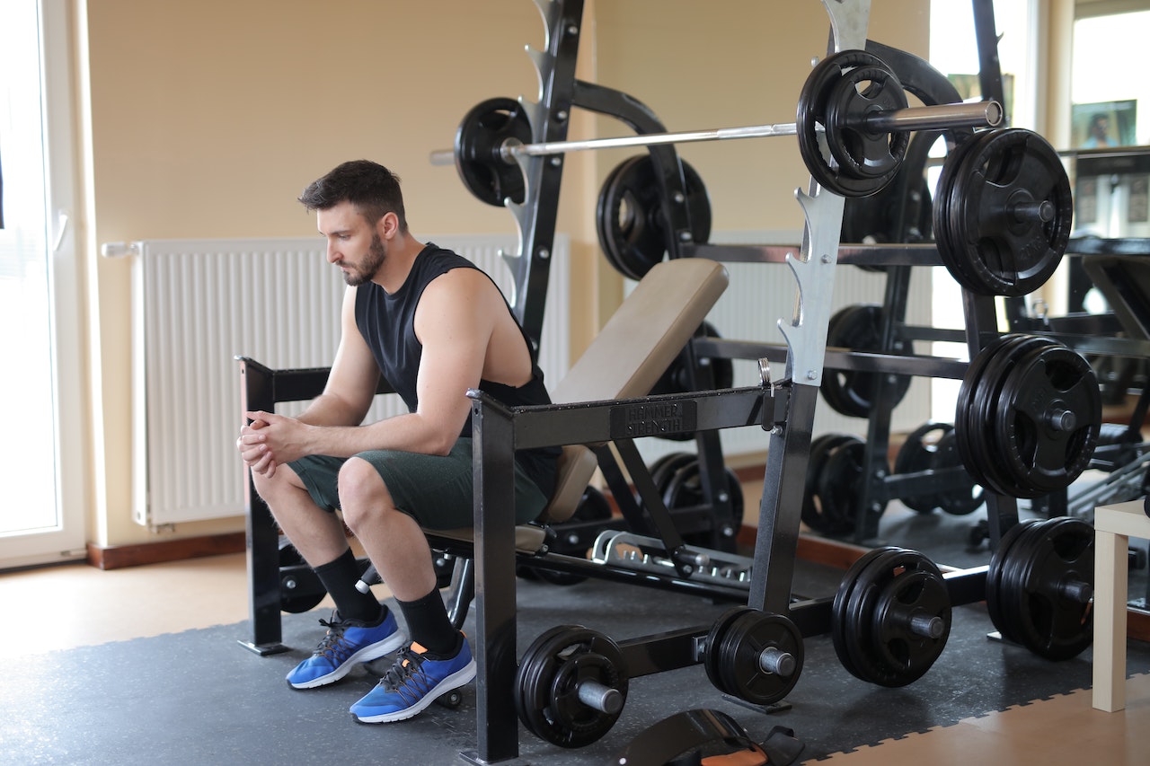 A man wearing a black tank top, gray shorts, and blue rubber shoes while sitting on a gym equipment