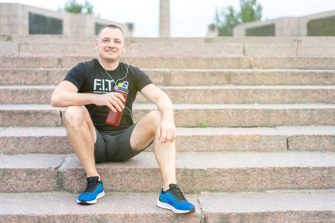 Man sitting on the steps outdoors while holding a red water tumbler and listening to his ear phones