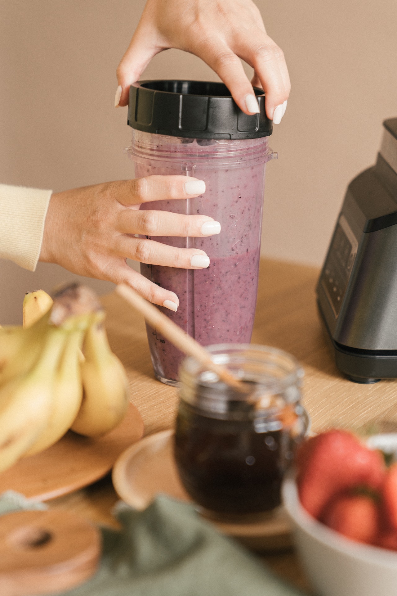 A person holding the black lid of a clear tumbler with mixed fruits near bananas, honey, and strawberry on top of a brown wooden table